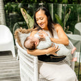 Load image into Gallery viewer, A mother bottle-feeding her child while the child sits comfortably in the HipSurfer Baby Carrier. The carrier supports comfortable feeding and offers ergonomic seating for the child.