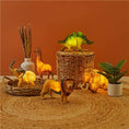Load image into Gallery viewer, Dinosaur Night Light placed on a wicker basket, surrounded by toy animals including a giraffe, lion, elephant, and mammoth.